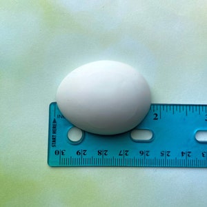May include: A white ceramic egg-shaped object measuring approximately 1 3/4 inches in length, placed on top of a blue ruler.