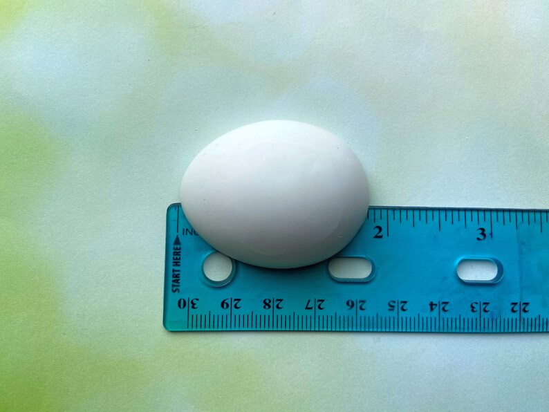 May include: A white ceramic egg-shaped object measuring approximately 1 3/4 inches in length, placed on top of a blue ruler.
