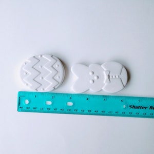 May include: Two white ceramic Easter-themed ornaments. One is an egg with a chevron pattern and the other is a bunny with a bow tie. The ornaments are approximately 2 inches in length.