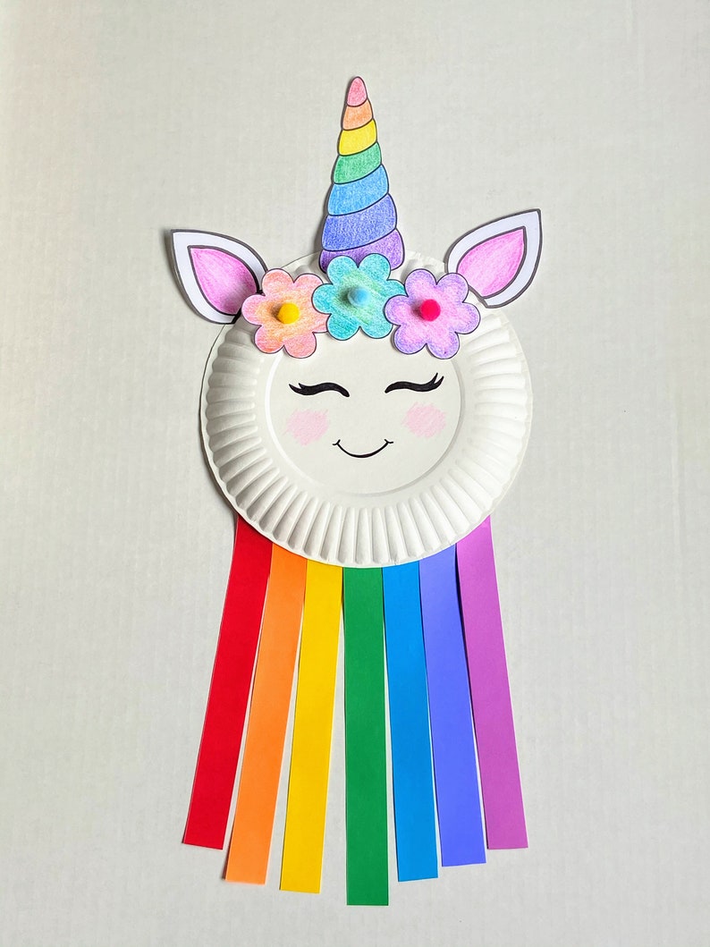 Example of finished unicorn paper plate craft. A face is drawn on a paper plate and a unicorn horn and ears glued on with flowers. Strips of colored paper hang from the bottom in a rainbow.
