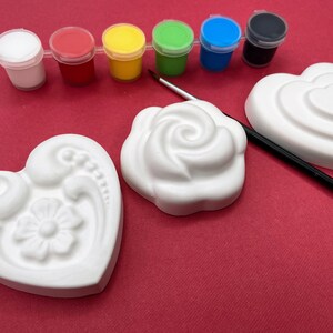 May include: A set of white ceramic heart-shaped ornaments with different designs, including a rose, a heart, and a floral pattern. The ornaments are ready to be painted with the included set of six paint pots in various colors, including white, red, yellow, green, blue, and black.