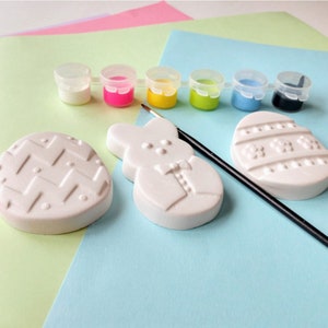 May include: A set of Easter-themed ceramic ornaments for painting, including a bunny, an egg with a chevron pattern, and an egg with a floral design. The set also includes six small paint pots in white, pink, yellow, green, blue, and black, and a paintbrush.