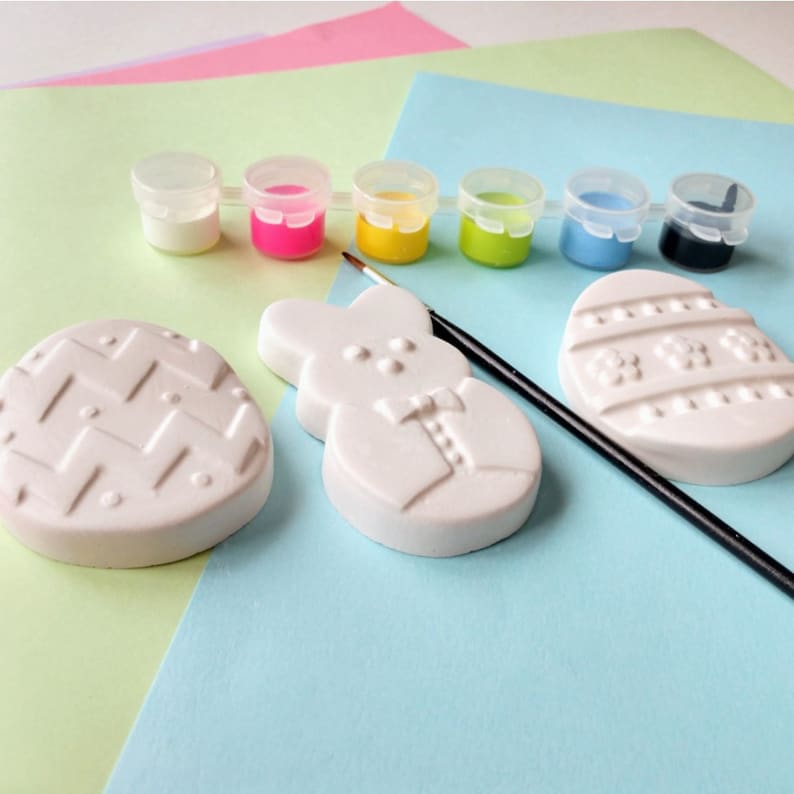 May include: A set of Easter-themed ceramic ornaments for painting, including a bunny, an egg with a chevron pattern, and an egg with a floral design. The set also includes six small paint pots in white, pink, yellow, green, blue, and black, and a paintbrush.