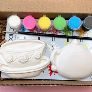 May include: A craft kit for painting a teacup and teapot. The kit includes two white ceramic pieces, six paint pots in various colors, and a paintbrush. The paint pots are labeled with the colors: white, pink, yellow, green, blue, and black.