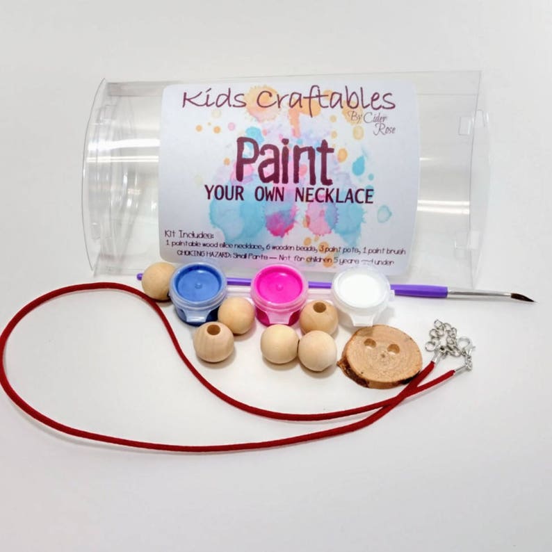 May include: A craft kit for making a necklace. The kit includes a red cord, wooden beads, paint pots, a paintbrush, and a wooden pendant. The kit is packaged in a clear plastic container with a label that reads "Kids Craftables Paint Your Own Necklace".