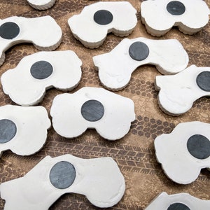 May include: A collection of white ceramic monster truck magnets with black magnet inserts. The magnets are arranged in a random pattern on a brown surface.