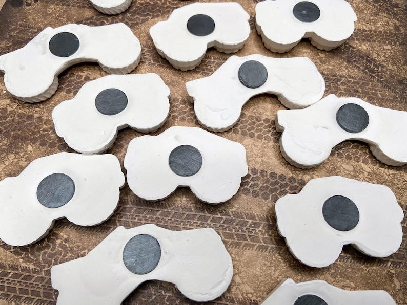 May include: A collection of white ceramic monster truck magnets with black magnet inserts. The magnets are arranged in a random pattern on a brown surface.