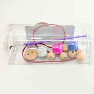 May include: A clear plastic container filled with craft supplies for making jewelry. The container includes wooden beads, a button, paint pots in white, pink, and blue, a paintbrush, and a red cord.