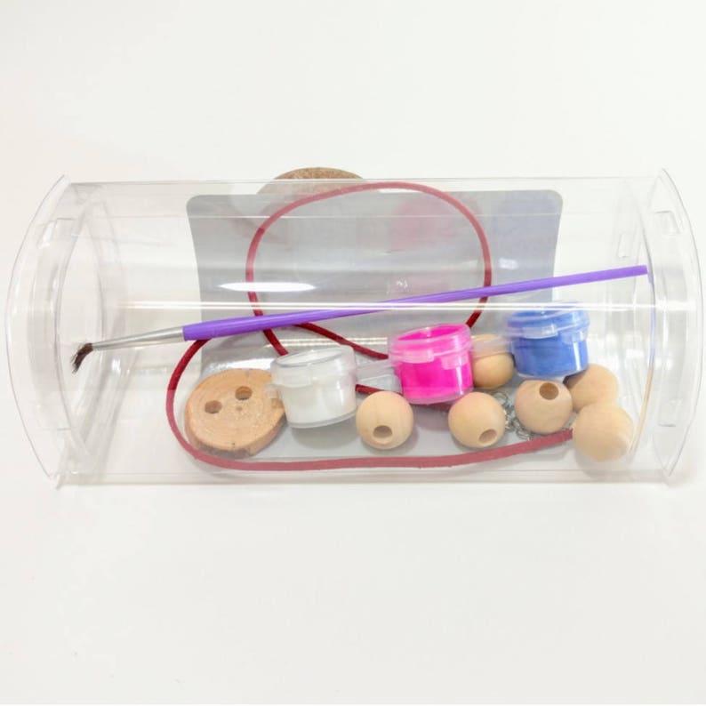 May include: A clear plastic container filled with craft supplies for making jewelry. The container includes wooden beads, a button, paint pots in white, pink, and blue, a paintbrush, and a red cord.