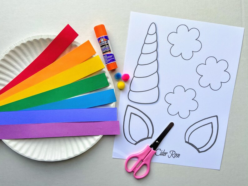 Items for the unicorn paper plate craft. One paper plate, seven colored strips of paper, glue, 3 pom poms, scissors and a cardstock age with a unicorn horn, two ears and three flower shapes to color and cut out.