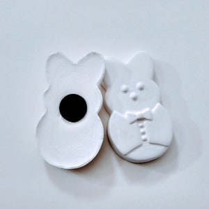 May include: Two white ceramic bunny figurines. One bunny has a round hole in the center and the other bunny has a bow tie and button details.