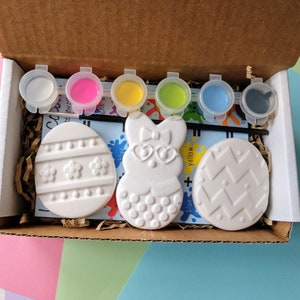 May include: A craft kit with three white ceramic Easter-themed ornaments, including an egg with stripes, an egg with a zig-zag pattern, and a bunny with a bow. The kit also includes six small paint pots in white, pink, yellow, green, blue, and black.