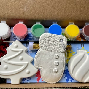 May include: A Christmas craft kit with six paint pots in white, red, green, blue, yellow, and brown. The kit includes three ceramic ornaments: a Christmas tree, a mitten, and an ornament ball. The ornaments are ready to be painted.
