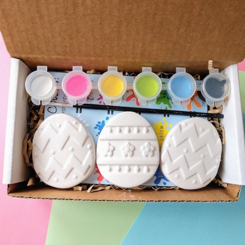 May include: A set of six paint pots with different colors, including blue, green, yellow, pink, white, and black. The paint pots are arranged in a row on a brown cardboard surface. There are three white ceramic Easter eggs with different designs on them.
