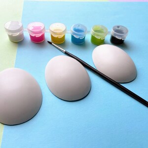 May include: Three white ceramic eggs, a paintbrush, and six small paint pots with lids. The paint pots are filled with white, pink, yellow, blue, green, and black paint.