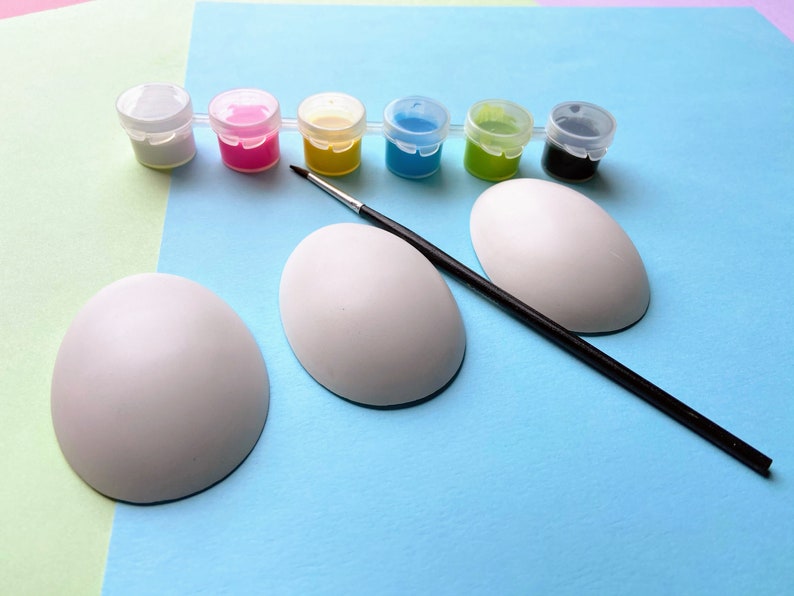 May include: Three white ceramic eggs, a paintbrush, and six small paint pots with lids. The paint pots are filled with white, pink, yellow, blue, green, and black paint.