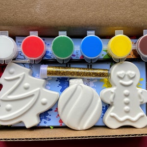 Puede incluir: Un kit de cerámica para pintar de temática navideña con adornos de cerámica blanca, un hombre de jengibre y un árbol de Navidad. El kit incluye seis botes de pintura en rojo, blanco, verde, azul, amarillo y marrón, un frasco de purpurina dorada y un pincel.