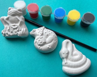 Puede incluir: Un kit de cerámica para pintar con tres figuras de animales: una rana, un camaleón y una serpiente. El kit incluye seis pequeños botes de pintura en blanco, rojo, verde, azul, amarillo y negro, y un pincel negro.