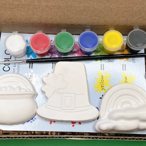 May include: A craft kit with three white ceramic pieces: a pot of gold, a leprechaun hat, and a rainbow. The kit includes six small paint pots in white, red, green, blue, yellow, and black, and a paintbrush.