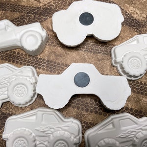 May include: Six white ceramic monster truck figurines with black magnet backs. The trucks have detailed wheels and some have flames painted on the sides.