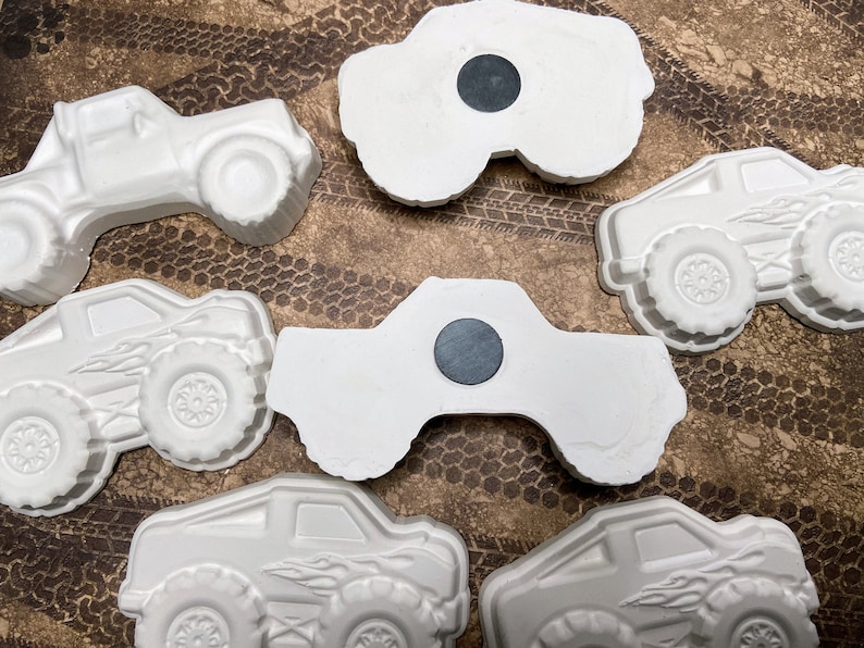 May include: Six white ceramic monster truck figurines with black magnet backs. The trucks have detailed wheels and some have flames painted on the sides.