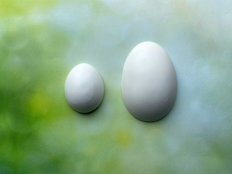 May include: Two white ceramic eggs, one larger than the other, on a green and blue background.