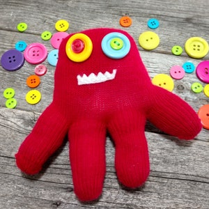 May include: A red knitted mitten with a white stitched mouth and two large eyes made from colorful buttons. The mitten is surrounded by colorful buttons on a wooden surface.