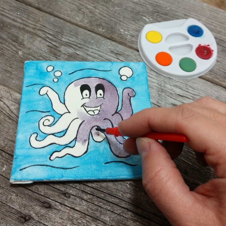 K&ouml;nnte beinhalten: Ein wei&szlig;er und lilafarbener Cartoon-Oktopus mit einem l&auml;chelnden Gesicht ist auf eine blaue Leinwand gemalt. Eine Hand h&auml;lt einen roten Pinsel und malt den Oktopus.