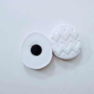 May include: Two white ceramic Easter eggs, one plain and one with a zig-zag pattern. The plain egg has a black circle in the center.