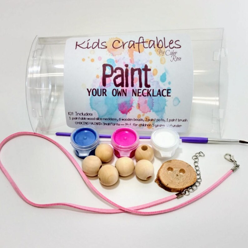 May include: A craft kit for making a necklace. The kit includes a pink cord, six wooden beads, a wooden pendant, three small paint pots, a paint brush, and the text "Kids Craftables Paint Your Own Necklace".