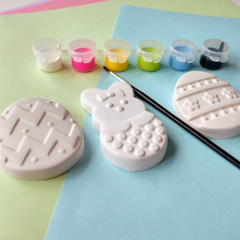May include: A set of Easter-themed paint-your-own ceramic ornaments. The set includes a bunny, an egg, and a basket. The ornaments are white and ready to be painted. The set also includes six small pots of paint in various colors.