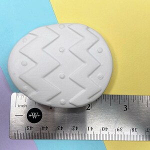 May include: White ceramic Easter egg with a chevron pattern and small dots. The egg is approximately 2 inches long and is shown next to a ruler.