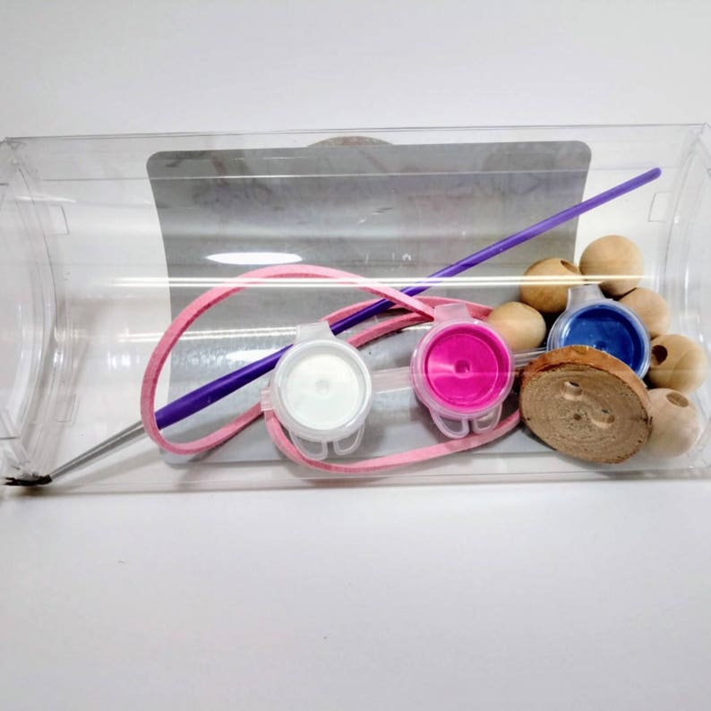 May include: A clear plastic container with a purple paintbrush, pink elastic, white, pink, and blue paint pots, wooden beads, and a wooden disc.