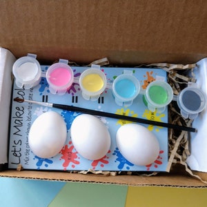 May include: A craft kit for painting eggs with six small paint pots in white, pink, yellow, blue, green, and gray. The kit includes three white ceramic eggs and a black paintbrush. The kit's packaging has the text "Let's Make Col." and instructions to mix the colors.