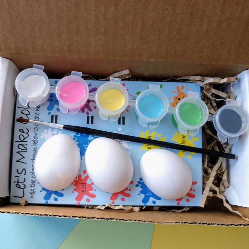 May include: A craft kit for painting eggs with six small paint pots in white, pink, yellow, blue, green, and gray. The kit includes three white ceramic eggs and a black paintbrush. The kit's packaging has the text "Let's Make Col." and instructions to mix the colors.