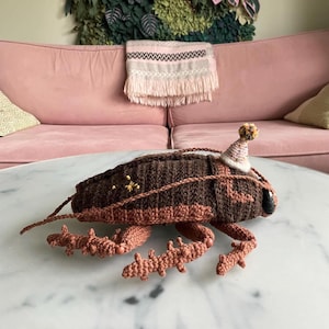Peut inclure: Un sac fait à la main en forme d'insecte crocheté, dans des tons de marron et de beige, orné d'un petit chapeau de fête. Le sac présente des pattes et des antennes détaillées, et est décoré de petites décorations jaunes. Le sac est posé sur une table en marbre.
