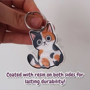 May include: Calico cat keychain with large eyes, sitting on a white background. The keychain is coated with resin on both sides for durability.