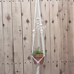 May include: Two macrame plant hangers with wooden beads hanging from a wooden fence. Each hanger has a small potted plant inside.