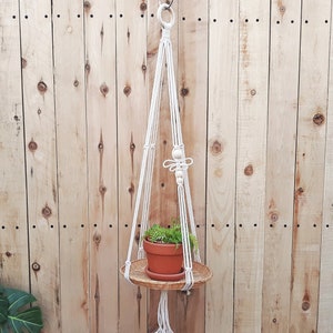 May include: A white macrame plant hanger with a wooden ring and a small potted plant. The hanger is hanging from a hook on a wooden wall.