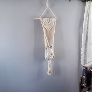 May include: A macrame plant hanger with a heart-shaped design. The hanger is made of natural cotton cord and has a wooden dowel at the top. A glass jar is hanging in the center of the hanger.