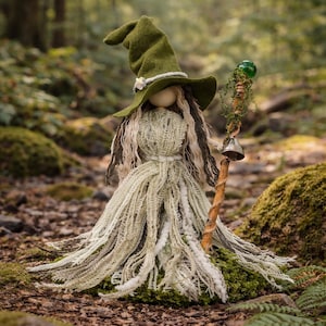 May include: A doll wearing a pointed olive green felt hat and a dress made of layered, fringed light green and white fabric. The doll has long, dark hair and holds a wooden staff topped with a green glass sphere and a small metal bell.