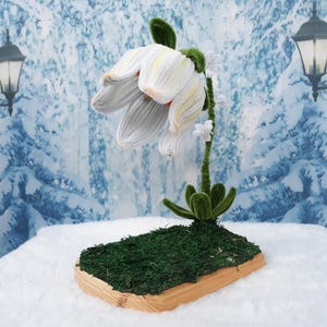 May include: A handcrafted, white and yellow flower sculpture with detailed petals and a green stem. The flower is mounted on a wooden base covered in green moss. The background features a winter scene with snow-covered trees and street lamps.