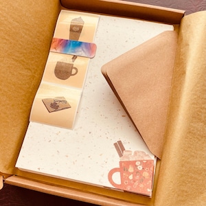 Könnte beinhalten: Ein Set aus braunem Papierbriefpapier mit einer weißen, gesprenkelten Oberfläche. Das Set enthält einen Stapel Briefpapier, einen braunen Umschlag und vier Aufkleber mit Illustrationen von Kaffee, einem Buch und einer Tasse heißer Schokolade.
