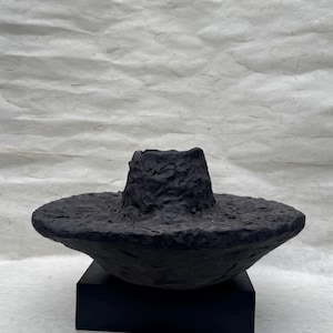May include: A dark, textured sculpture, possibly a hat or bowl, with a broad brim and a central, cylindrical form. The artwork is presented on a black, rectangular base, against a textured, off-white backdrop.