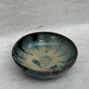 May include: A handcrafted ceramic bowl with a circular form. The bowl displays a mix of blue, green, and beige hues, with darker detailing around the edge. The inside has a speckled design, and the outside is smooth.