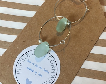 May include: A pair of silver hoop earrings with a light green sea glass charm hanging from each hoop. The earrings are displayed on a brown paper bag with a white circle label that reads "Pebble Cornwall Cornish sea glass silver jewellery Handmade by Terre".
