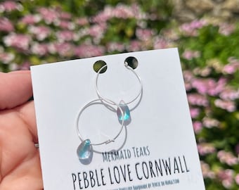 May include: Sterling silver hoop earrings with a teardrop-shaped iridescent blue gemstone. The earrings are labelled "Mermaid Tears Pebble Love Cornwall".