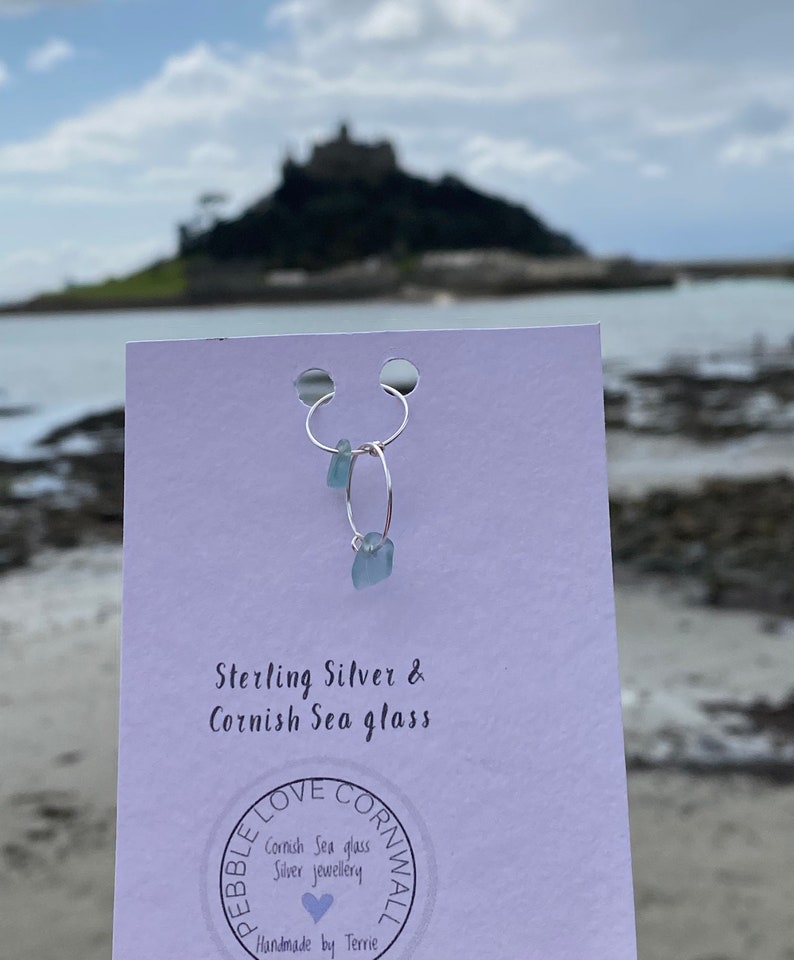 May include: Sterling silver hoop earrings with a light blue sea glass charm. The earrings are displayed on a white card with the text "Sterling Silver & Cornish Sea glass" and a logo that says "Pebble Love Cornwall, Cornish Sea glass Silver Jewellery, Handmade by Terrie".
