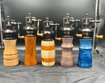 May include: Five wooden pepper grinders with metal mechanisms and wooden handles. The grinders have different wood finishes, including light oak, cherry, and blue-stained wood. The metal components have a bronze finish.