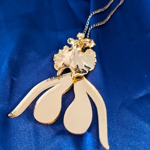 Op de afbeelding: Een gouden ketting met een grote, abstracte hanger. De hanger is gevormd als een gestileerd menselijk figuur met een vloeiende rok en een gespiegelde oppervlak.